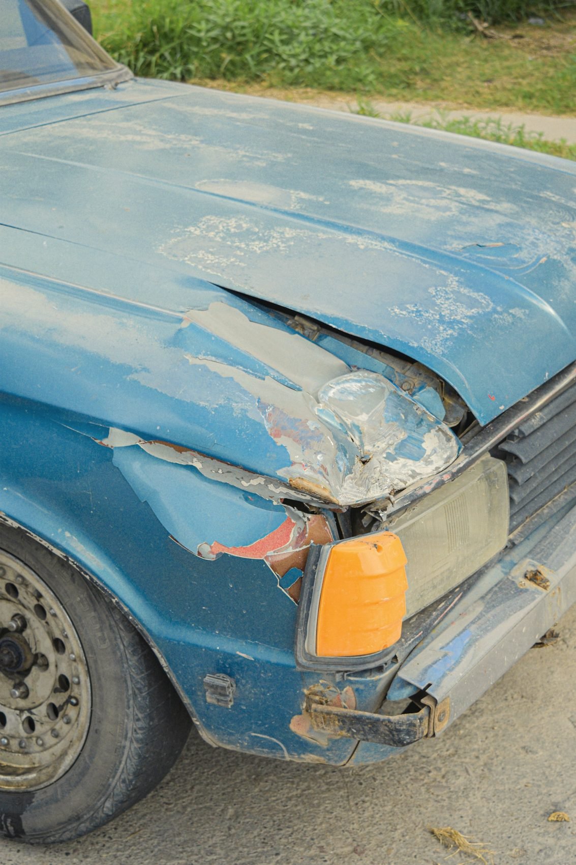 The dented front corner of an old blue car with flaking paint and a cracked headlight