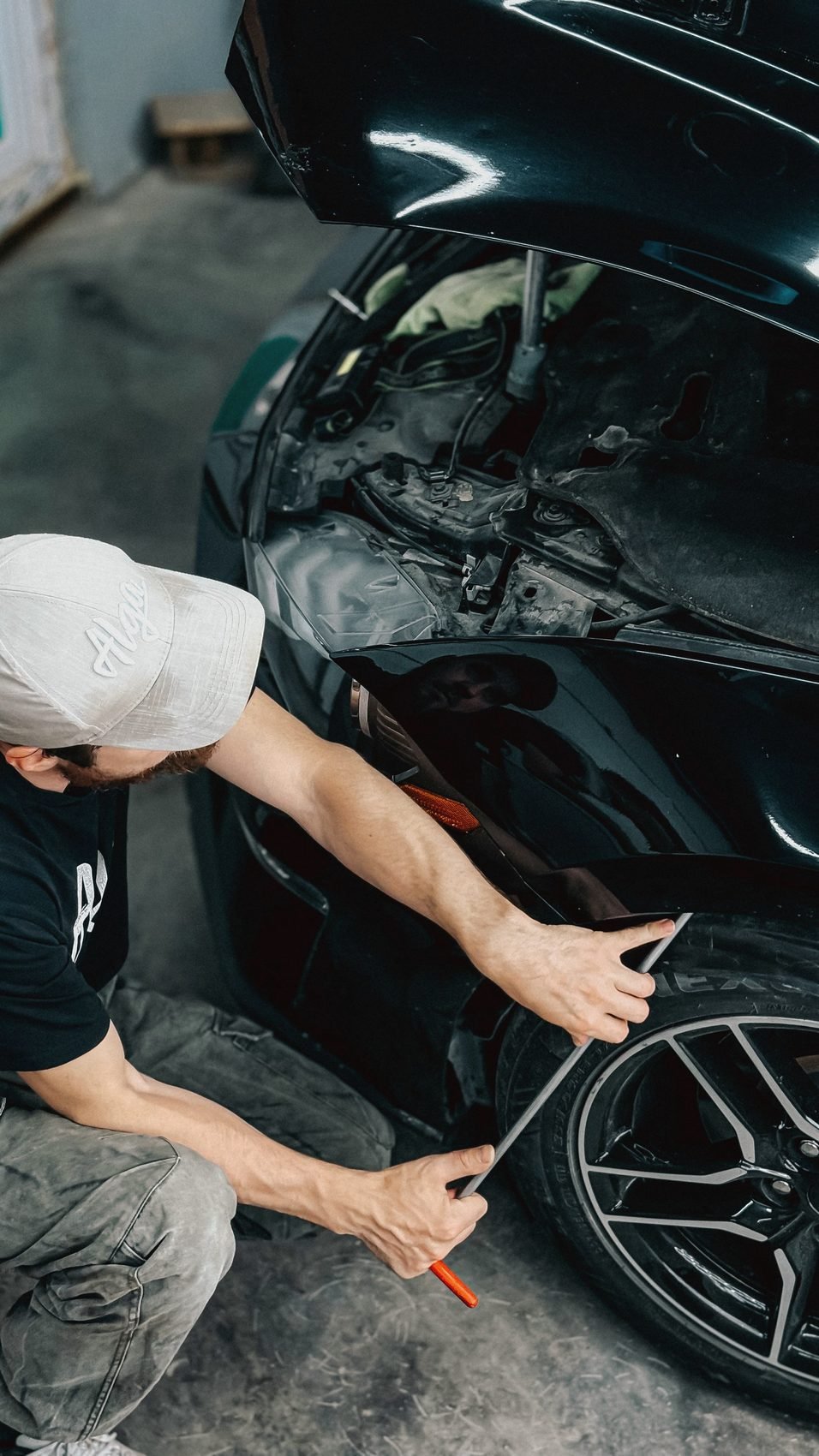 A person in a white cap crouched at the front wheel arch of a dark car with a hand tool