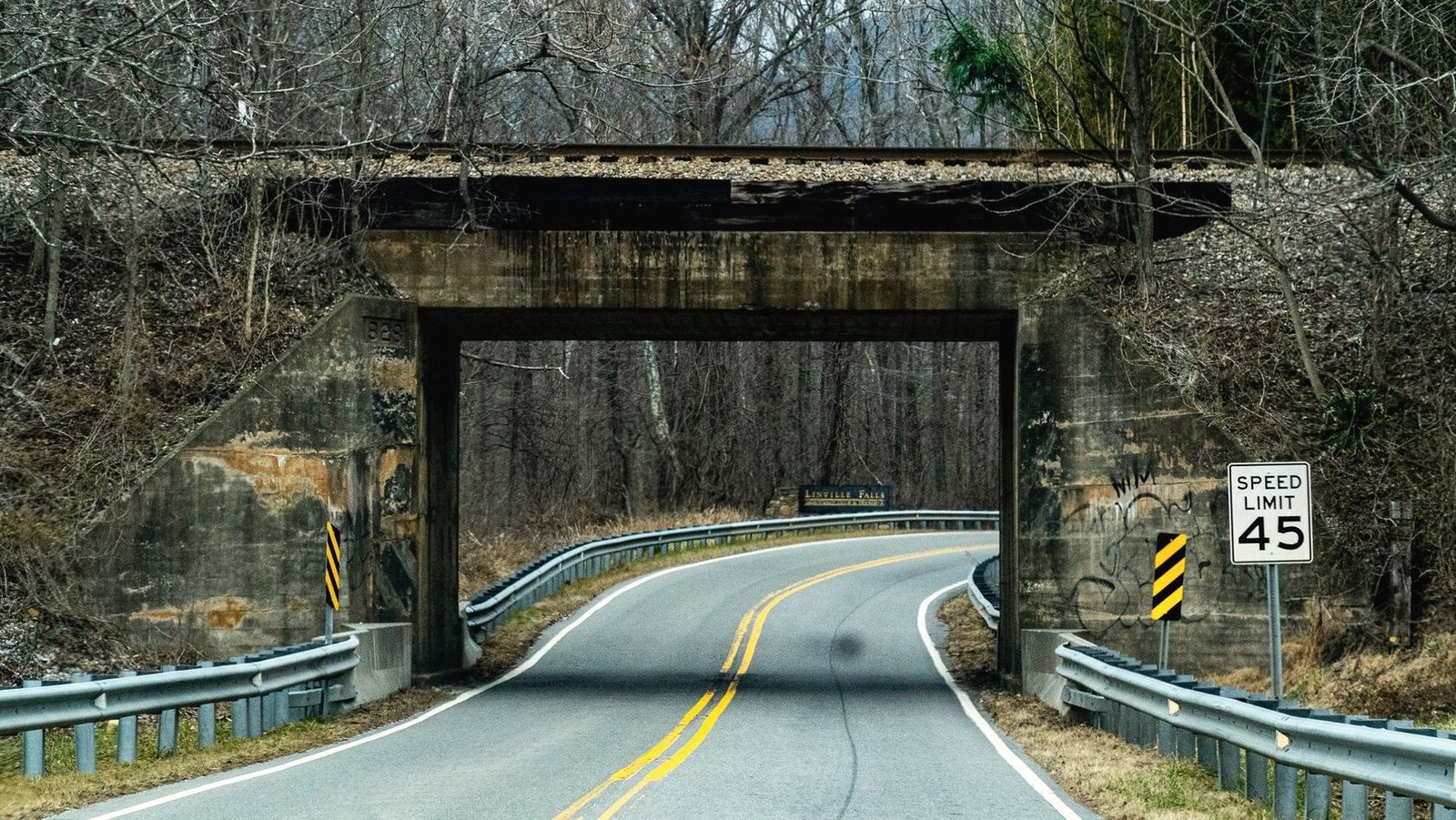 A two-lane road with a double yellow centre line curving under an old railroad overpass, with a 45 mile per hour speed limit sign