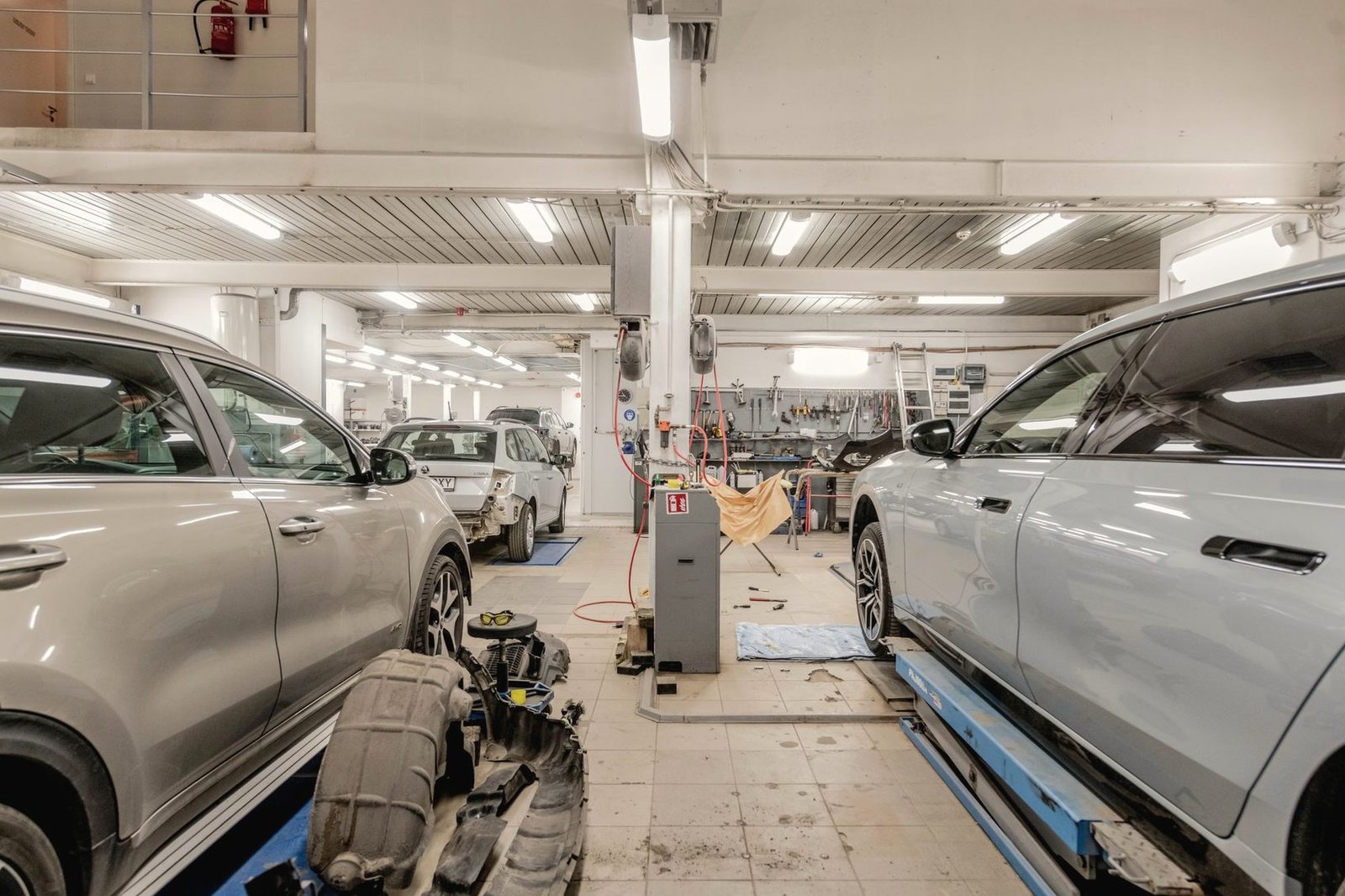 The floor of a repair shop with two silver cars raised on blue racks and a removed bumper on the ground